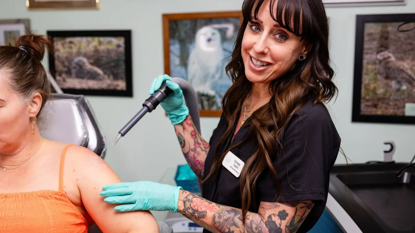 A woman with long brown hair, full sleeve tattoos, wearing gloves and a black shirt, in a clinic with owl pictures on the wall. The owner of the clinic chilling her client with a cryo machine before preforming a tattoo removal treatment.