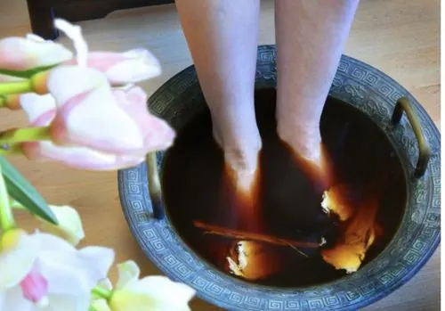 Person soaking feet in a bowl of dark herbal foot soak, with flowers nearby.