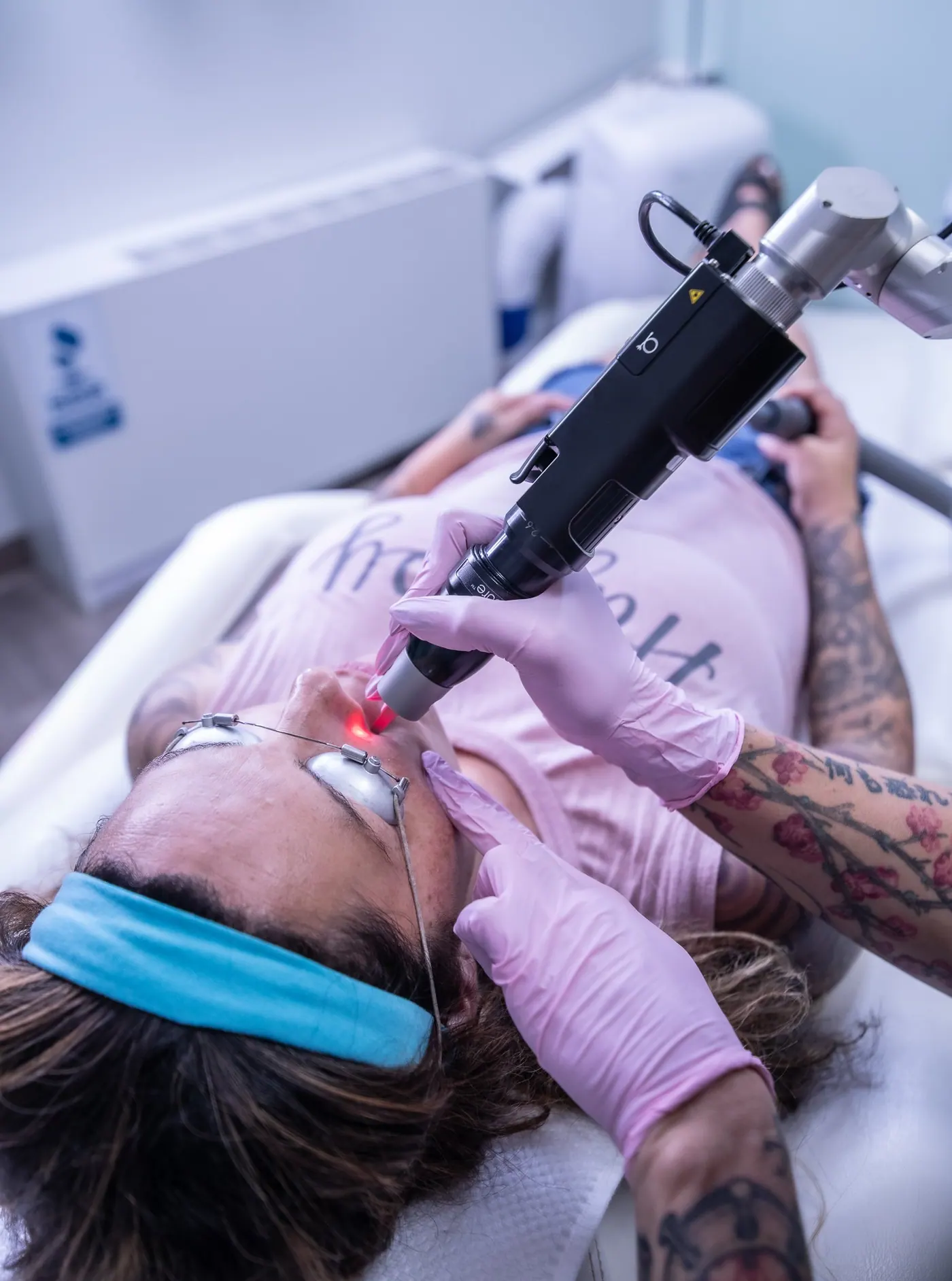 Tattooed woman getting a laser tattoo removal procedure on her upper lip at a spa or clinic, wearing protective eye gear, with a technician using a laser device.