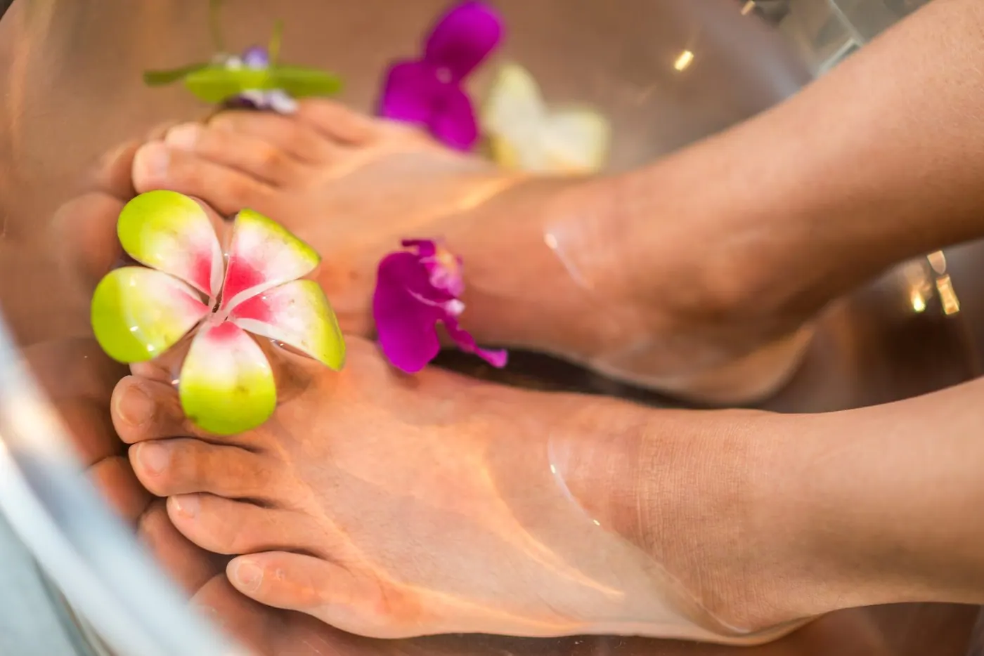 Feet soaking in a foot bath with pink and purple flowers and a green and white flower on top.